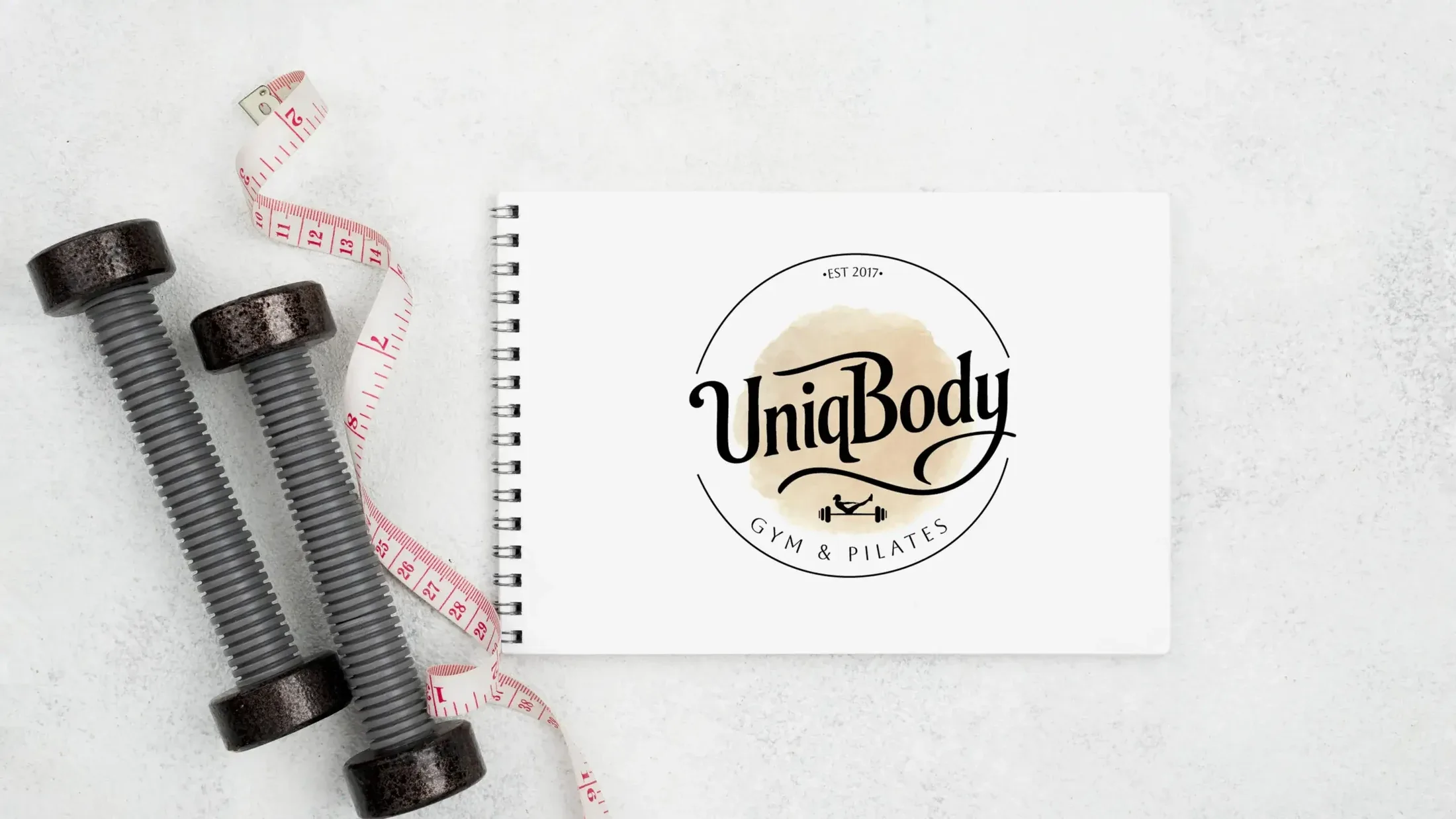 Logo UniqBody w otwartym notatniku obok hantli i centymetra krawieckiego